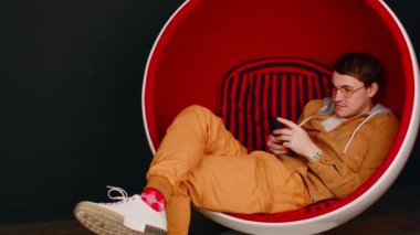 Adam yumurta koltuğunda akıllı telefona bakıyor. Beyaz ve kırmızı yumurta şekilli koltukta oturan erkek hipster siyah arka planda cep telefonuyla sosyal medyada mesajlaşıyor..