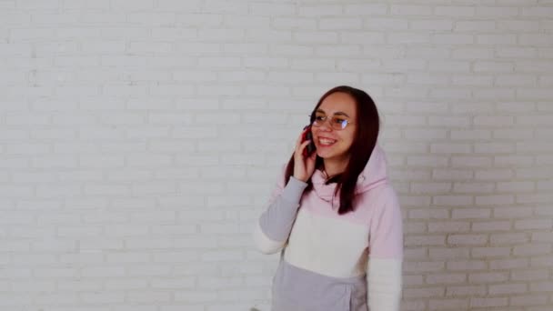 안경을 쓴 젊은 여성은 스마트폰으로 흰색 벽돌 벽 가까이에 서서 말을 한다. 휴대 전화로 하는 예쁜 여성 강연. — 비디오