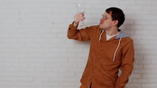 Homem de óculos com copo de vinho na parede de tijolo branco. O jovem macho acaba o vinho. Homem irritado treme vidro vazio sobre a boca. — Vídeo de Stock