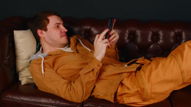 Człowiek przeglądający smartfony na kanapie. Skupiony mężczyzna w ubraniu casual SMS na komórkę podczas odpoczynku na wygodnej skórzanej kanapie w pokoju na czarnym tle — Wideo stockowe