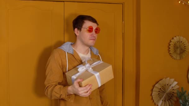 Young man in bright glasses with wrapped gift box standing near doors in room. Stylish male looking away and holding present. — Stockvideo