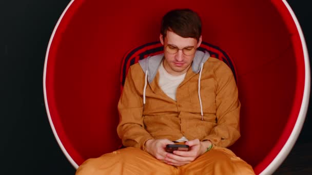 Muž prohledávající smartphone v křesle s vejci. Muž hipster sedí v bílém a červeném vejci ve tvaru křesla a poselství na sociálních sítích přes mobil na černém pozadí. — Stock video
