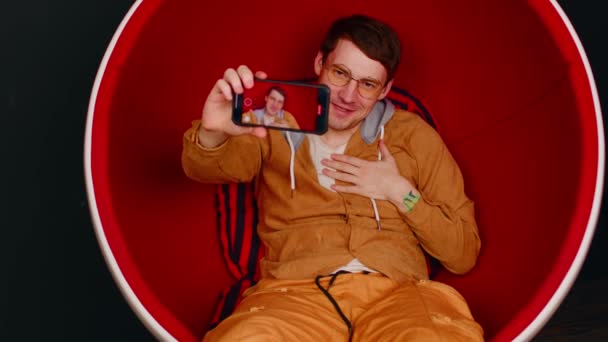 Koltuğunda selfie çeken neşeli bir adam. Modern ışık stüdyosunda kırmızı koltukta otururken şık gözlüklü bir erkek akıllı telefonuyla kendi portresini çekiyor. — Stok video