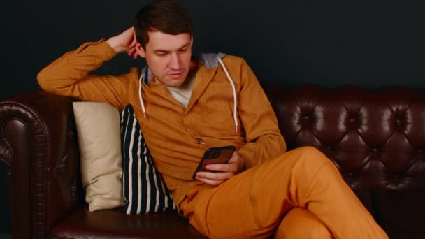 年轻人坐在沙发上靠在枕头上浏览手机。使用智能手机释放男性，并在业余时间休息。靠近点. — 图库视频影像