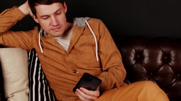 Молодий чоловік переглядає мобільний телефон, сидячи на дивані, спираючись на подушки. Розслаблений чоловік використовує смартфон і відпочиває у вільний час. крупним планом . — стокове відео