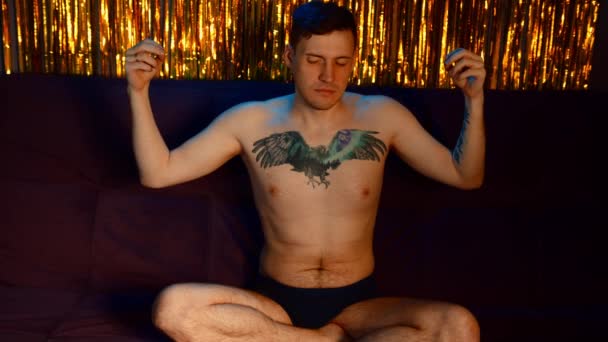 Naked man meditating on sofa in dark room against golden tinsel. Serene male with closed eyes sitting in easy pose keeping hands in zen gesture and trying to relax. — Stockvideo