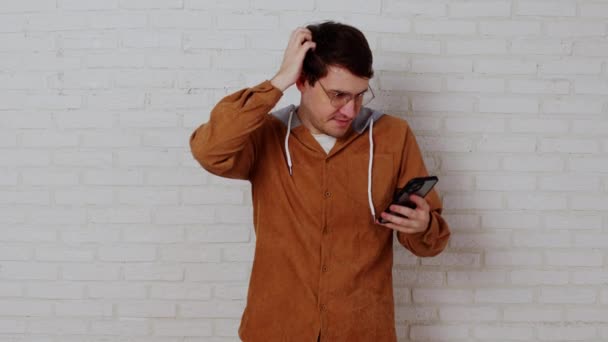 Προσεκτικός άνθρωπος ψάχνει στο κινητό τηλέφωνο στο παρασκήνιο του λευκού τοίχο τούβλο. Pensive αρσενικό σε γυαλιά ξύσιμο του κεφαλιού και τη χρήση smartphone. — Αρχείο Βίντεο