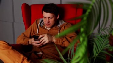 Akıllı telefonlu bir adam koltukta oturuyor. Koyu renk elbiseli, koltukta oturan ve akıllı telefonuyla mesajlaşan ciddi bir genç erkek.
