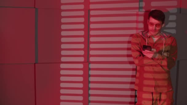 Άνθρωπος με γυαλιά περιήγηση στο κινητό τηλέφωνο, στέκεται κοντά στον τοίχο με σκιές της jalousie και χώρο για κείμενο. Κομψό αρσενικό χρησιμοποιώντας smartphone σε σκοτεινό δωμάτιο με κόκκινο φως που πέφτει και σκιά περσίδες. — Αρχείο Βίντεο