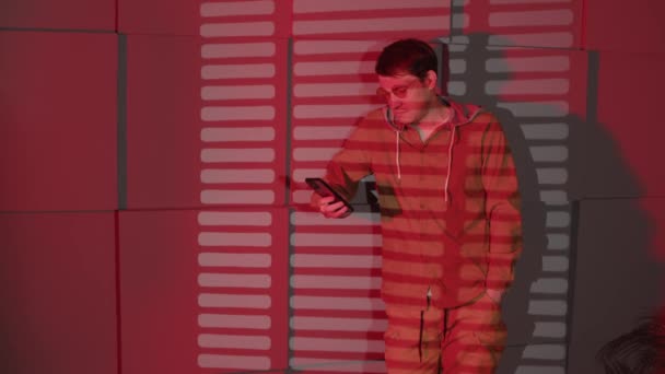 Man in glasses browsing mobile phone, standing near wall with shadows of jalousie and space for text. Stylish male using smartphone in dark room with falling red light and shadow blinds. — Stockvideo