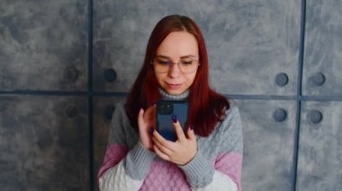 Ciddi, siyah saçlı, sıradan giyimli, akıllı telefon kullanan genç bir kadın. Güneş gözlüklü bir kadın beton duvarın yanında cep telefonuna bakıyor.