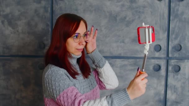 Young woman in glasses with mobile phone standing near wall. Positive female in knitted sweater shoots video on smartphone. Happy lady conducts live broadcast with friends on cellphone. — Stockvideo
