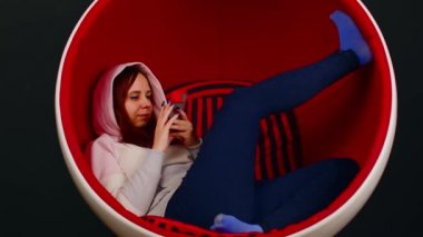 Cep telefonlu genç bir kadın siyah arka planda top sandalyesinde oturuyor. Kadın akıllı telefona göz atıyor ve modern koltukta dinleniyor..