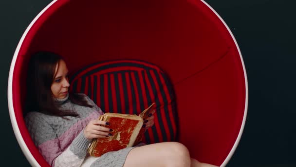 Young woman reads old book, sitting in ball chair. Female resting with shabby book in modern armchair. — Stockvideo