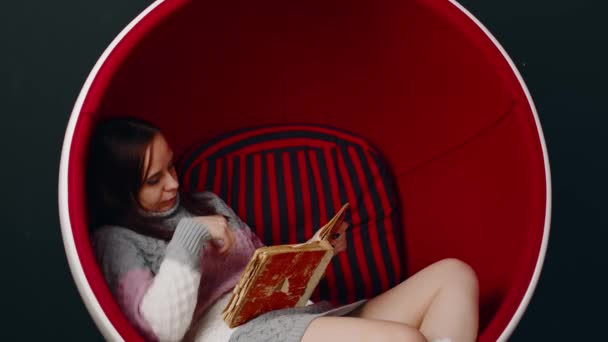 Una joven lee un libro viejo, sentada en una silla de baile. Reposo femenino con libro de mala calidad en sillón moderno. — Vídeo de stock