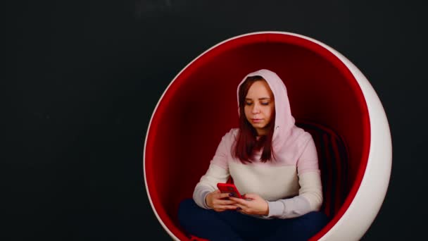 Mujer navegando smartphone en sillón de huevo. Mujer hipster sentada en un sillón con forma de huevo blanco y rojo y mensajería en las redes sociales a través del teléfono celular sobre fondo negro — Vídeo de stock