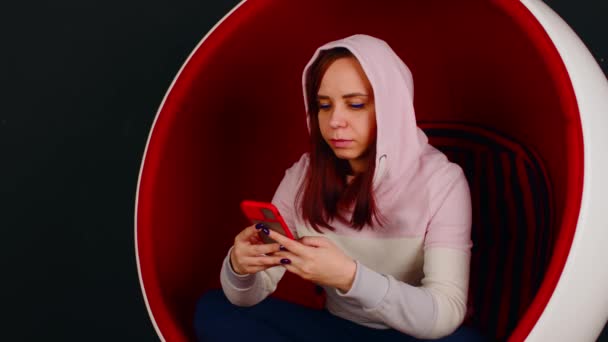 Ung kvinna med mobiltelefon sitter i bollstol på svart bakgrund. Kvinna bläddra smartphone och vila i modern fåtölj. — Stockvideo