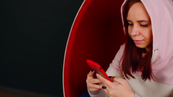 Een vrouw in een eierfauteuil. Vrouwelijke hipster zittend in witte en rode eivormige fauteuil en messaging op social media via mobiele telefoon op zwarte achtergrond — Stockvideo