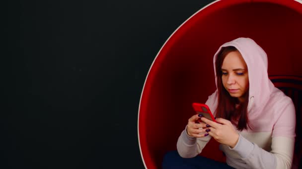 Женщина просматривает смартфон в яичном кресле. Женщина-хипстер сидит в белом и красном кресле в форме яйца и отправляет сообщения в социальных сетях через мобильный телефон на черном фоне — стоковое видео