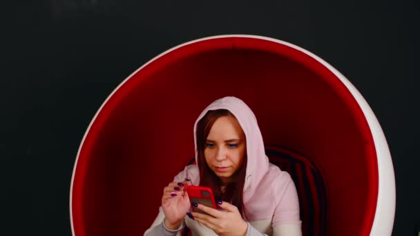 Kobieta przeglądająca smartfona w fotelu na jajka. Kobieta hipster siedzi w białym i czerwonym fotelu w kształcie jajka i wiadomości w mediach społecznościowych za pośrednictwem telefonu komórkowego na czarnym tle — Wideo stockowe