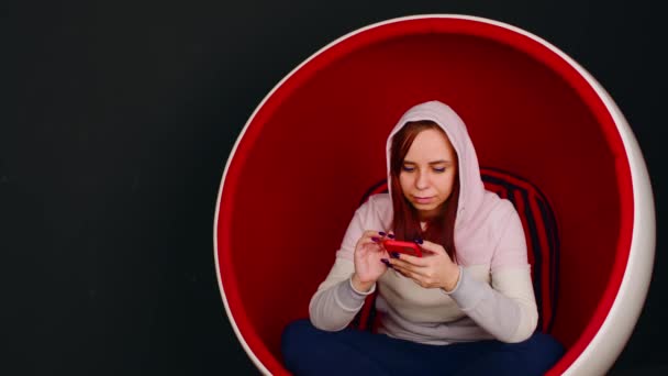 Женщина просматривает смартфон в яичном кресле. Женщина-хипстер сидит в белом и красном кресле в форме яйца и отправляет сообщения в социальных сетях через мобильный телефон на черном фоне — стоковое видео