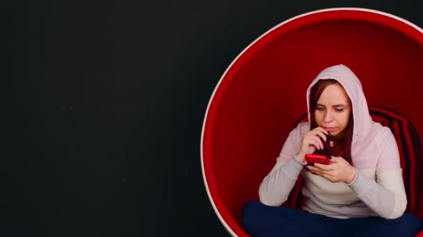 Fiatal nő mobiltelefonnal ül golyóálló székben, fekete háttérrel. Női böngészés okostelefon és pihenés a modern karosszékben. — Stock videók