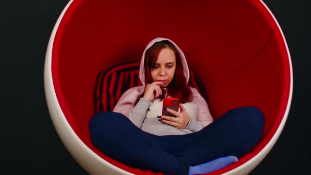 Een vrouw in een eierfauteuil. Vrouwelijke hipster zittend in witte en rode eivormige fauteuil en messaging op social media via mobiele telefoon op zwarte achtergrond — Stockvideo