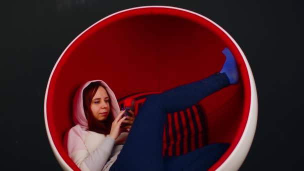 Young woman browsing mobile phone and sitting with her legs up in ball-shaped chair. Female in hood using smartphone and resting in modern armchair during free time. — Stockvideo