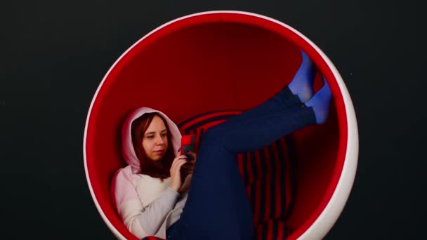 Woman browsing smartphone in egg armchair. Female hipster sitting in white and red egg shaped armchair and messaging on social media via cellphone on black background — 图库视频影像