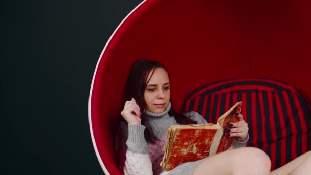 Young woman reads old book, sitting in ball chair. Female resting with shabby book in modern armchair. — Stockvideo