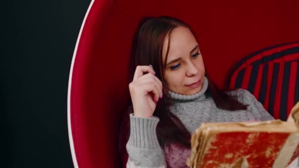 Young woman reads old book, sitting in ball chair. Female resting with shabby book in modern armchair. — Video Stock