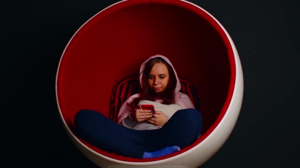 卵のアームチェアでスマートフォンを閲覧女性.白と赤の卵の形をしたアームチェアに座っている女性のヒップスターと黒の背景に携帯電話を介してソーシャルメディア上のメッセージ — ストック動画