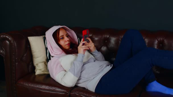 Kobieta przeglądająca smartfona na kanapie. Skupiona kobieta w luźnej bluzie SMS-y z kapturem na komórce podczas odpoczynku na wygodnej skórzanej kanapie w pokoju na czarnym tle — Wideo stockowe