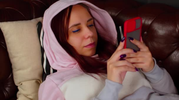 Kvinna bläddrar smartphone på soffan. Fokuserad kvinna i casual hoodie SMS på mobiltelefon samtidigt vilar på bekväm läder soffa i rummet på svart bakgrund — Stockvideo