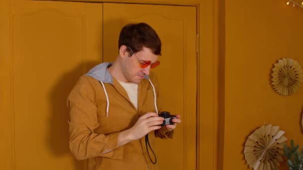 Młody mężczyzna w pomarańczowych okularach ze starym aparatem fotograficznym stoi na tle brązowych drzwi w pokoju. Stylowy mężczyzna uważa stary aparat fotograficzny i robi zdjęcia, patrząc w aparat. — Wideo stockowe