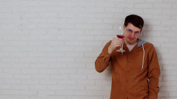 Jeune homme dans des verres avec verre à vin sur un mur de briques blanches. Homme détendu boit du vin rouge et regarde la caméra, debout près du mur blanc avec de l'espace pour votre texte. — Video