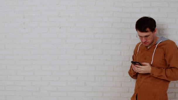 Młody człowiek w okularach przeglądający telefon komórkowy na białej ceglanej ścianie. Stylowy mężczyzna przy użyciu smartfona, stojący przy białej ścianie z miejscem na tekst. — Wideo stockowe