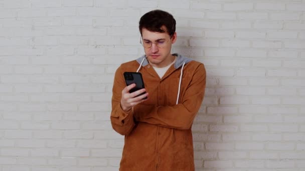 Young man in glasses browsing mobile phone on white brick wall. Stylish male using smartphone, standing near white wall. — Stock Video