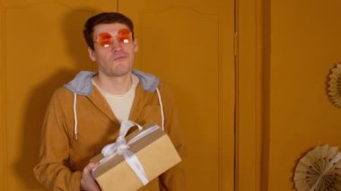 Paketli hediye kutusu olan parlak gözlüklü genç bir adam. Tarz sahibi erkek kameraya Noel hediyesi veriyor. Mutlu adam kameraya bakıyor, gülümsüyor ve kurdeleyle bağlanmış hediyeye uzanıyor..