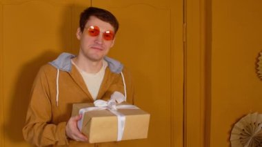 Paketli hediye kutusu olan parlak gözlüklü genç bir adam. Mutlu erkek kameraya bakıyor, gülümsüyor ve Noel hediyesini tutuyor..