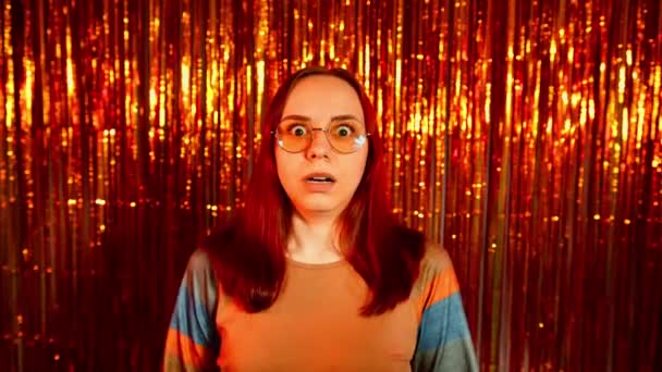 Shocked woman in glasses looking at camera on background of shiny tinsel. Surprised young female opens mouth, leaning hands against cheeks and shakes head. – Stock-video