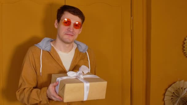 Νεαρός με φωτεινά γυαλιά και τυλιγμένο κουτί δώρου. Ευτυχισμένο αρσενικό κοιτάζει κάμερα, χαμογελώντας και κρατώντας χριστουγεννιάτικο δώρο. — Αρχείο Βίντεο