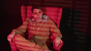 Dudaklarını ısıran düşünceli bir adam. Karanlık odada koltukta oturuyor. Düşen kırmızı ışık ve gölge panjurları var. Gözlüklü dalgın bir erkek hayatı yansıtıyor..