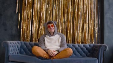 Young man in hood with ears and bright glasses sitting on sofa against wall with golden tinsel. Relaxed male sitting on couch leaning against back. Concept of rest and free time.