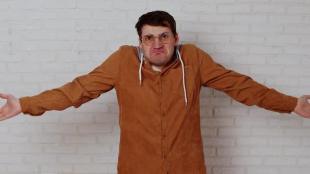 Young man in glasses spreading hands on background of white brick wall. Perplexed male shows gesture of not knowing, looking at camera. — Stock Video