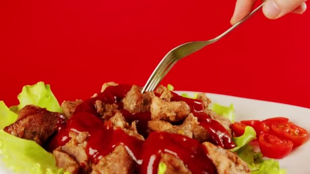 Una mano de hombre con un tenedor sumerge un trozo de carne en ketchup sobre un fondo rojo. El primer plano de las manos de las mujeres usando una cubertería, comiendo carne abundantemente vertida con salsa roja — Vídeos de Stock