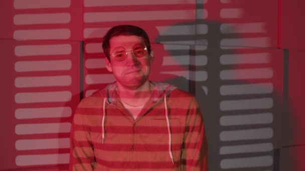 Positive man in glasses standing near wall in dark room with falling red light and shadow blinds. Handsome male looking at camera, smiling and blowing kisses. — Stock Video