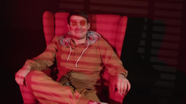 Junger Mann mit Brille sitzt in einem Sessel in einem dunklen Raum mit fallendem Rotlicht und Schattenjalousien. Positiver Mann blickt in die Kamera und macht Diktion-Übungen mit dem Mund. — Stockvideo