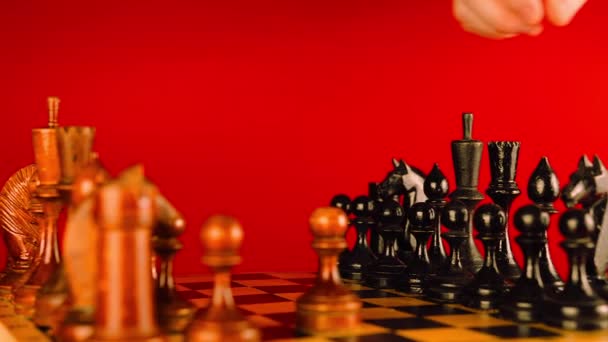 ボード上の白と黒の木製のチェスの作品を閉じます 男の手は赤い背景でチェス盤の上に黒い駒を動かす インテリジェントな 論理的かつ戦略的なゲームの概念 — ストック動画