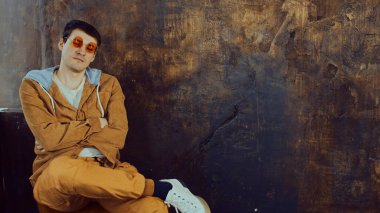 Parlak gözlüklü genç adam eski püskü kahverengi duvarın yanında oturuyor ve mesaj için yeri var. Rahat stil sahibi erkek kollarını kavuşturup kameraya bakar.